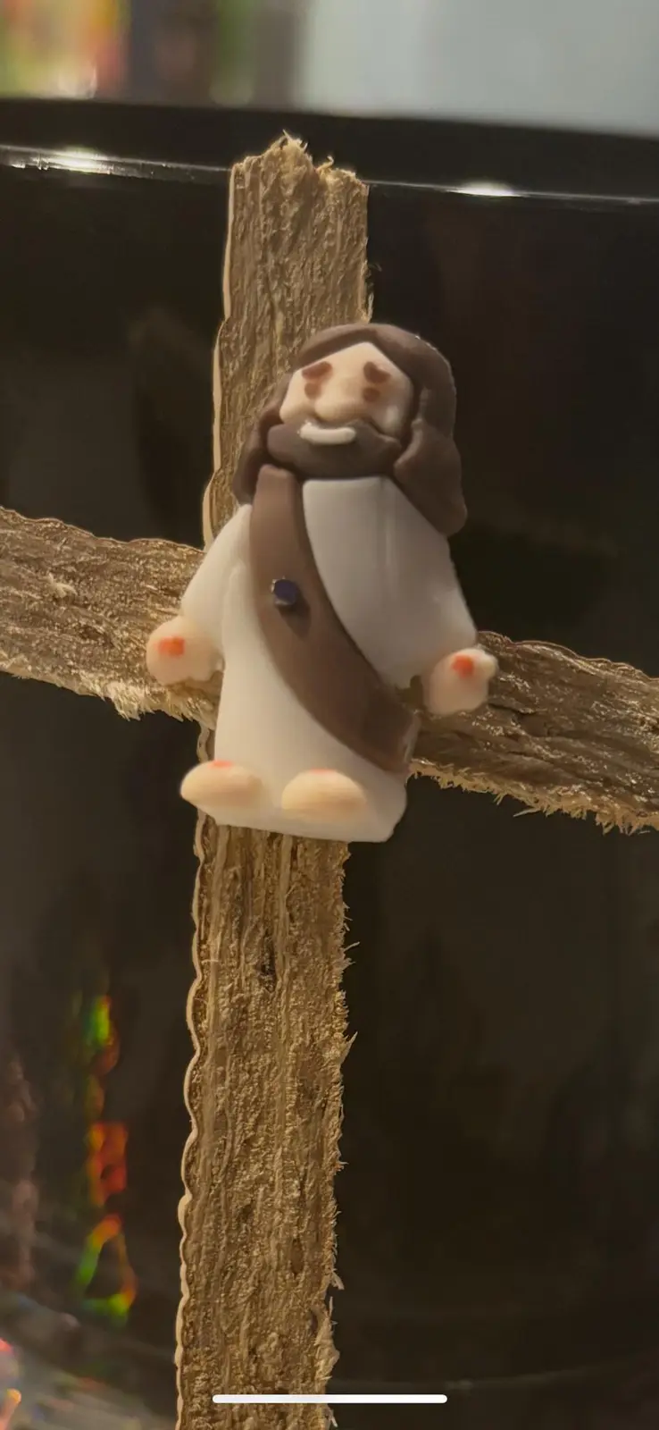 A small, stylized figurine of Jesus with a smiling face, brown hair and beard, and white robe is mounted on a rough, brown…