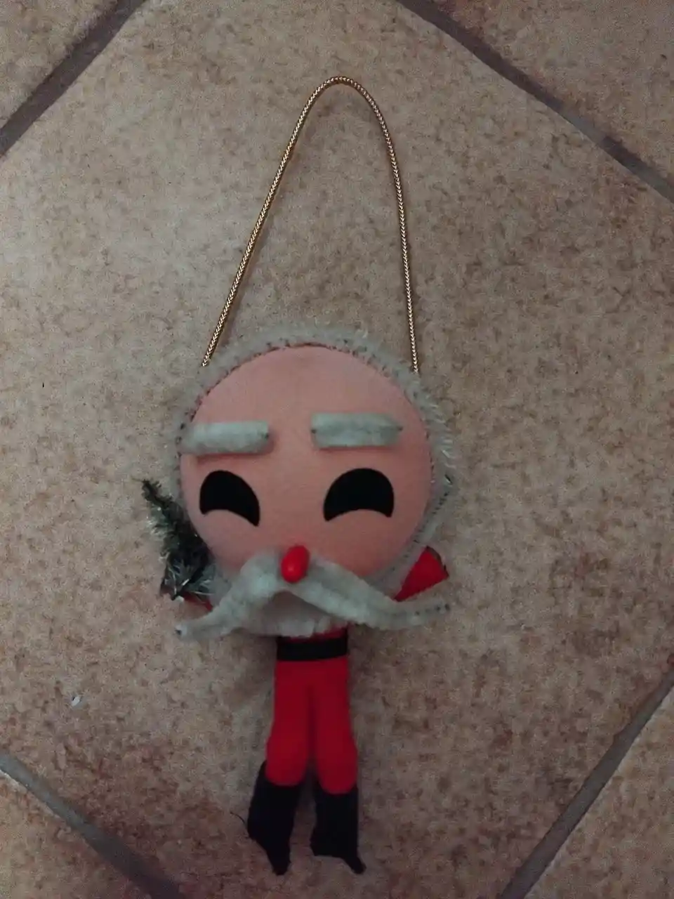 A handmade felt Santa Claus ornament with a pink face, white beard, red nose, and red suit lies on a tiled floor. It has a…