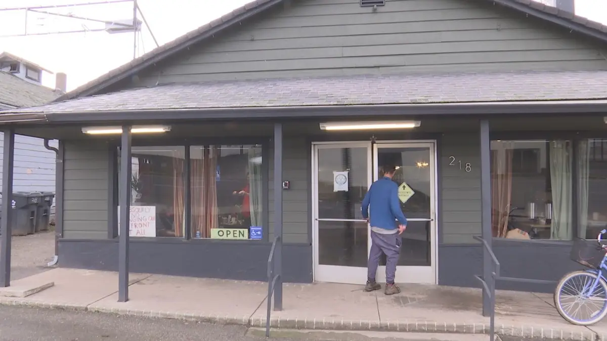A man in a blue shirt and gray pants stands facing the glass door of a small gray building with the number 218 on it…
