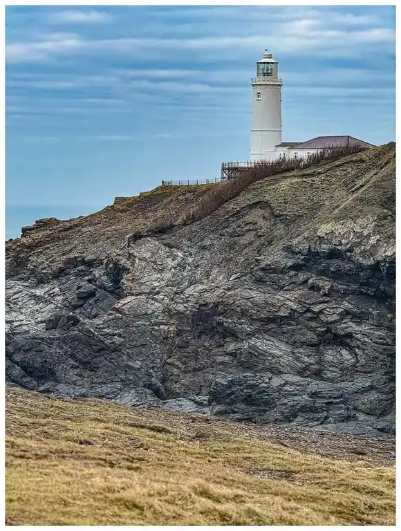 A white lighthouse stands atop a rugged, grassy cliff under a partly cloudy sky. The image features overlaid text reading…