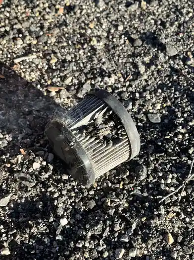 A dirty, clogged, and visibly damaged air filter lies on gravel, with smoke rising from it, overlaid with the text 'i'm not…