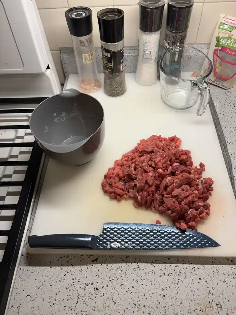 A kitchen counter shows a pile of freshly chopped raw meat on a white cutting board, with a large chef's knife resting in…