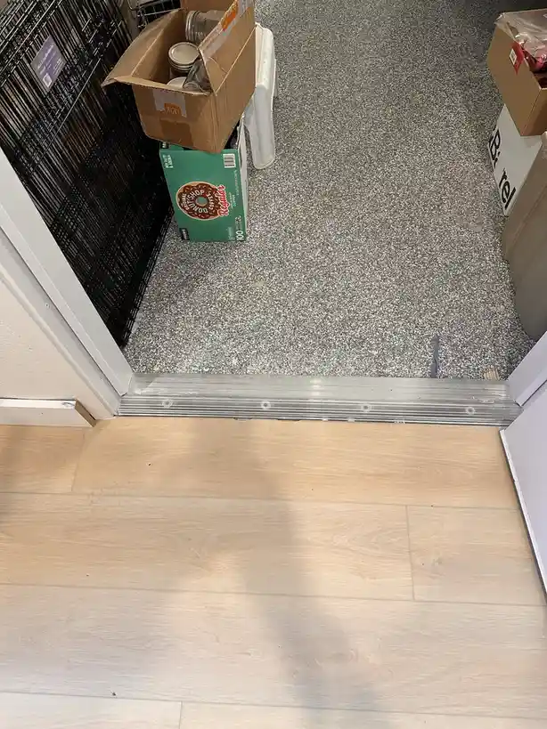 A photo shows a doorway with a light wood floor on the inside and a speckled gray floor on the outside, where a green box of…