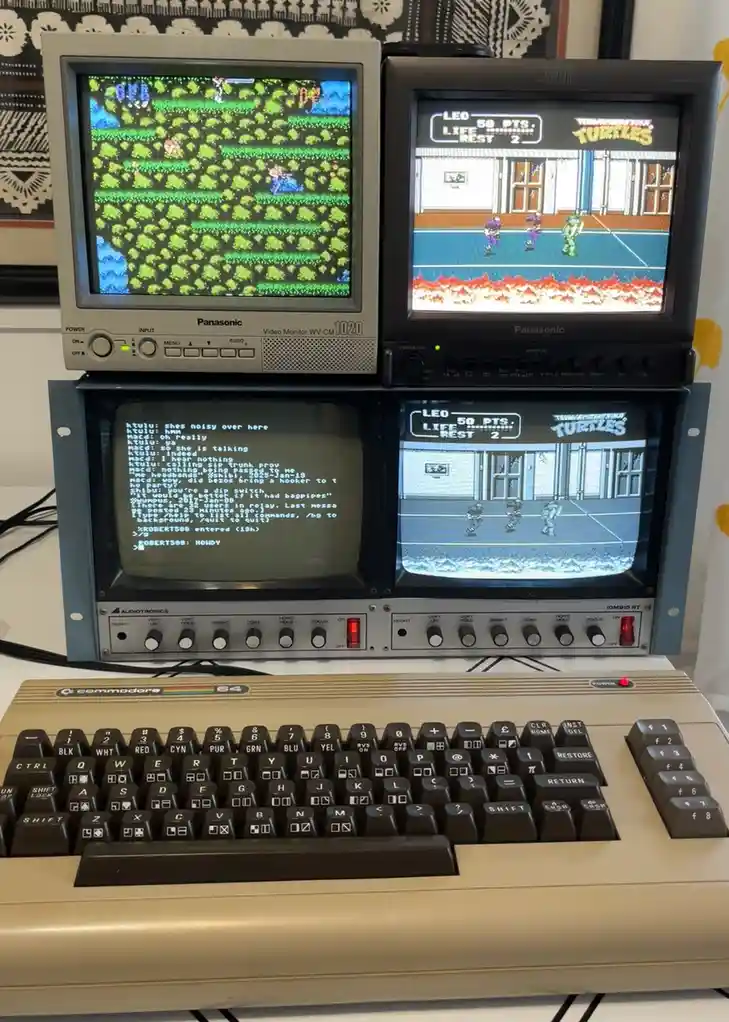 A vintage Commodore 64 computer with a beige keyboard sits in front of a rack holding four small Panasonic monitors. Two…