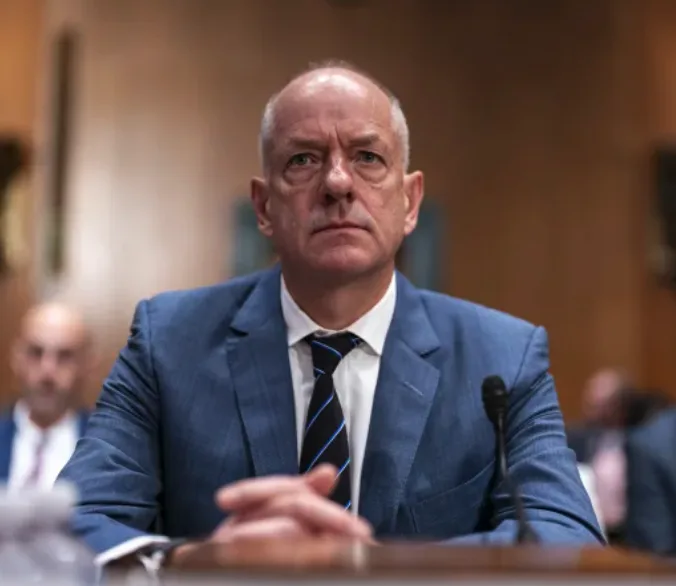 a man in a blue suit and striped tie sits at a wooden table with a microphone, looking intensely at the camera with a stern…
