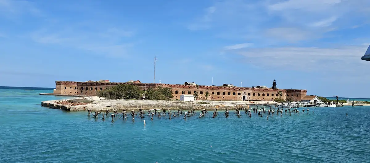 A wide shot of a large, historic brick fort on a small island surrounded by turquoise ocean water under a blue sky. In the…