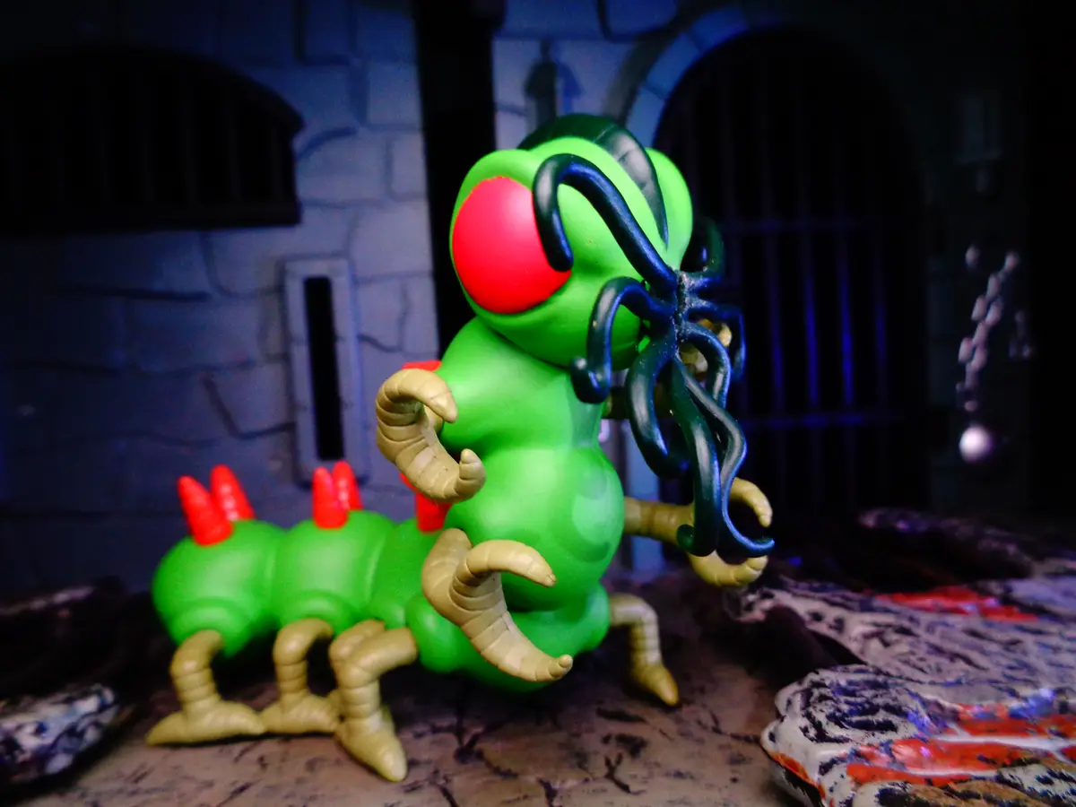 A green plastic caterpillar toy with red spikes and tan legs stands in a dimly lit, dungeon-like setting with stone walls…
