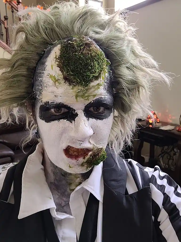 A person in a Beetlejuice costume with white face paint, black eye makeup, and a wild white wig, has green moss growing out…