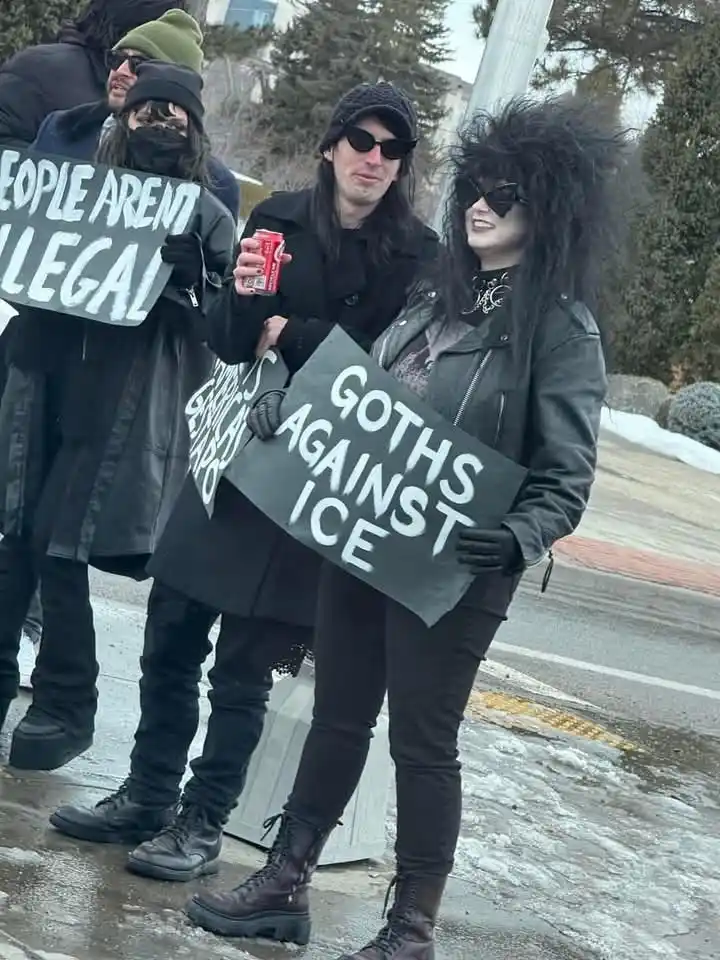 A group of people dressed in goth fashion, including black clothing, sunglasses, and dramatic hairstyles, are protesting…
