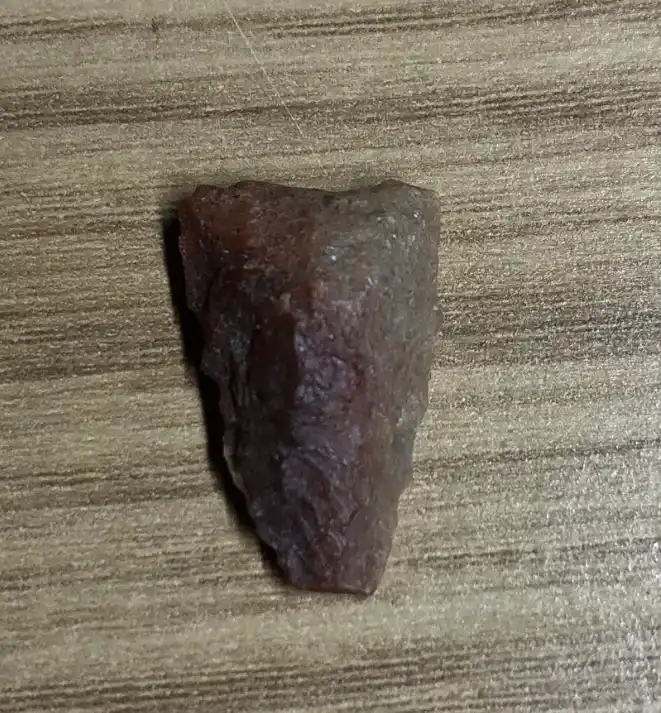 A small, dark brown, irregularly shaped stone with a rough, chipped surface rests on a light brown wooden surface with…