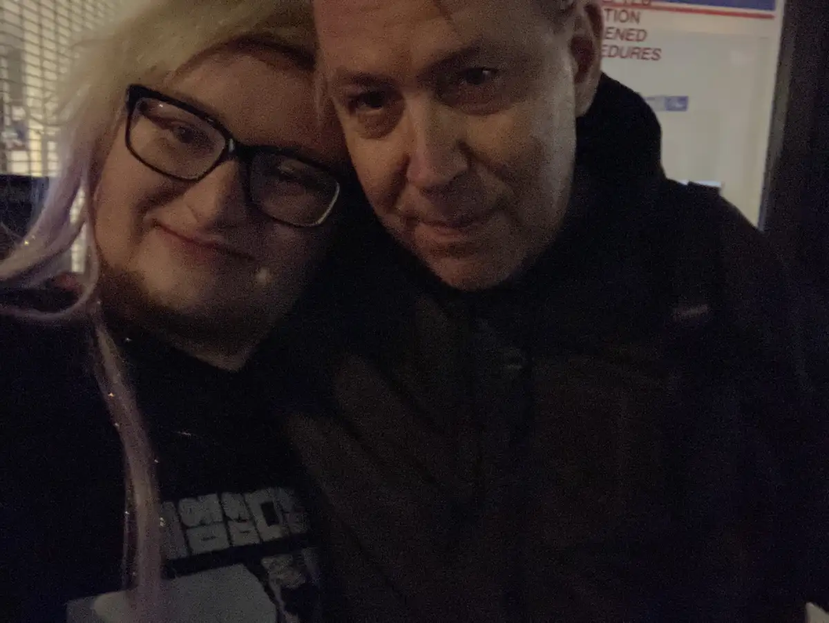 A selfie shows a woman with glasses and a blonde wig smiling while leaning against a man in a dark jacket who has a neutral…