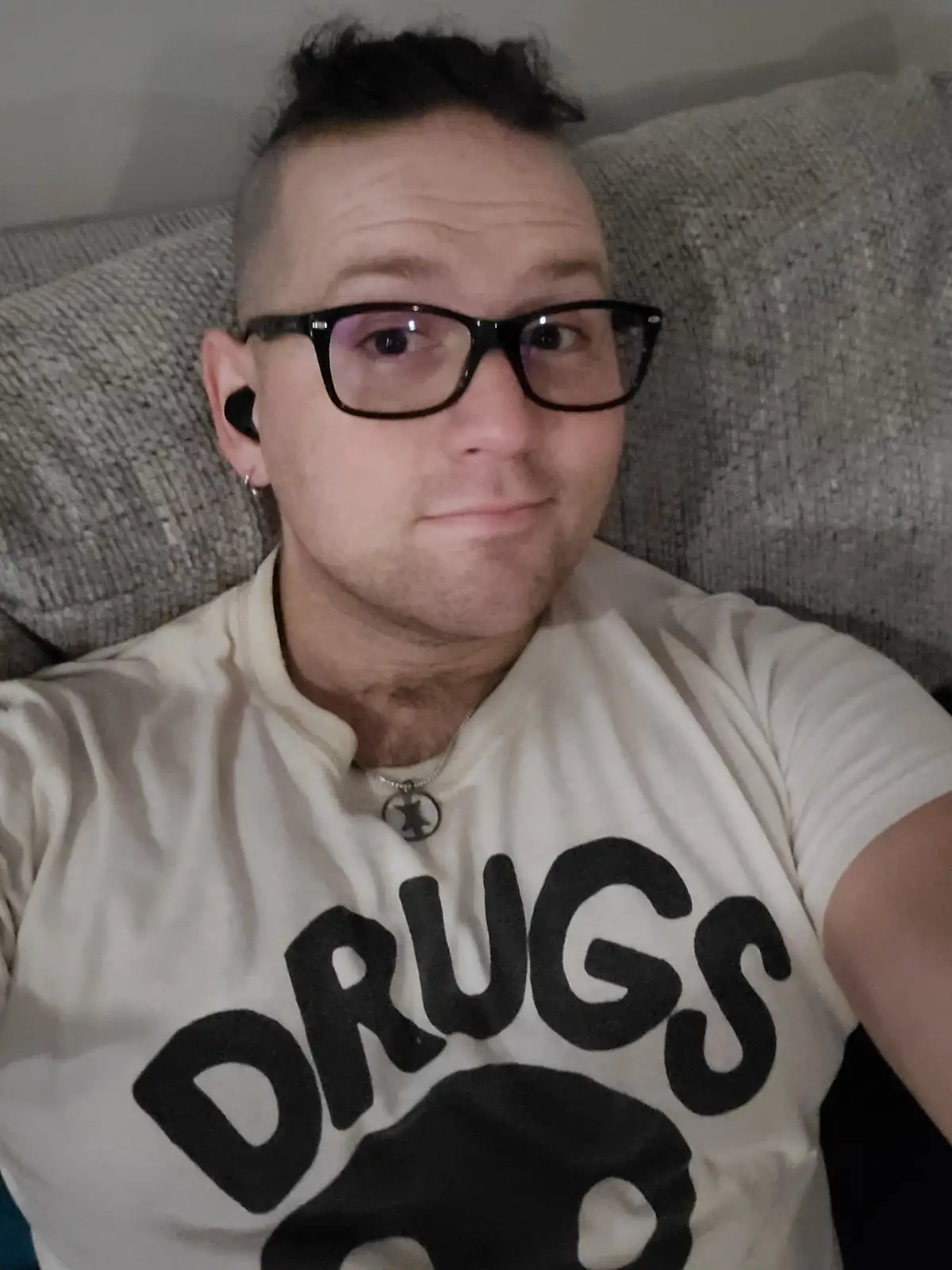 A man with glasses and a shaved head wearing a white t-shirt with the word 'DRUGS' in large black letters is lying on a gray…