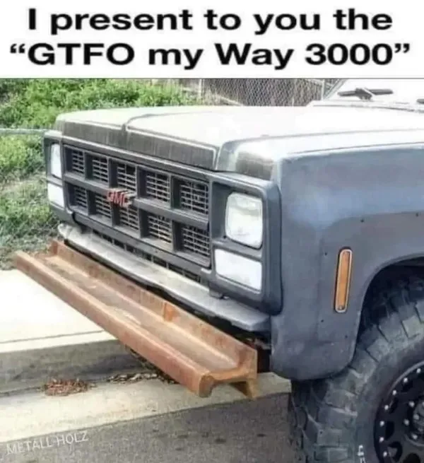 A rusty, modified GMC truck with a large, rusted metal beam bolted to its front bumper, protruding outward. The overlaid…