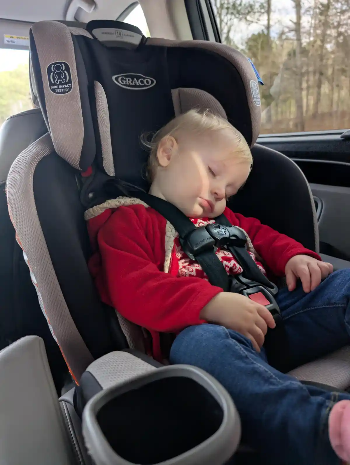 A young child with blonde hair is peacefully sleeping in a Graco car seat inside a vehicle, wearing a red fleece jacket and…