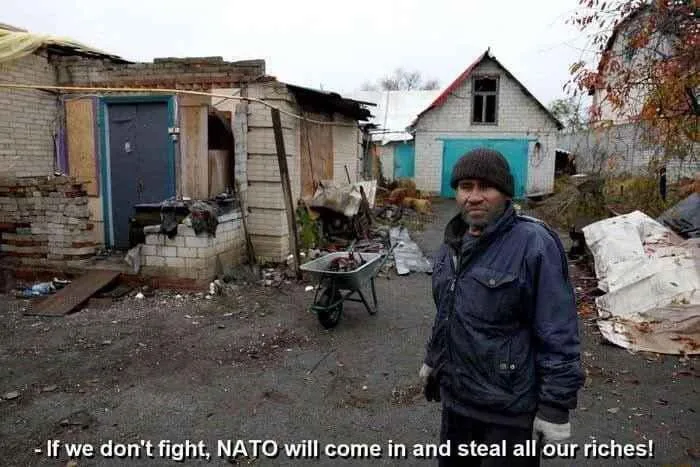 A man in a dark jacket and beanie stands in a dilapidated, debris-strewn yard with crumbling brick structures and a…