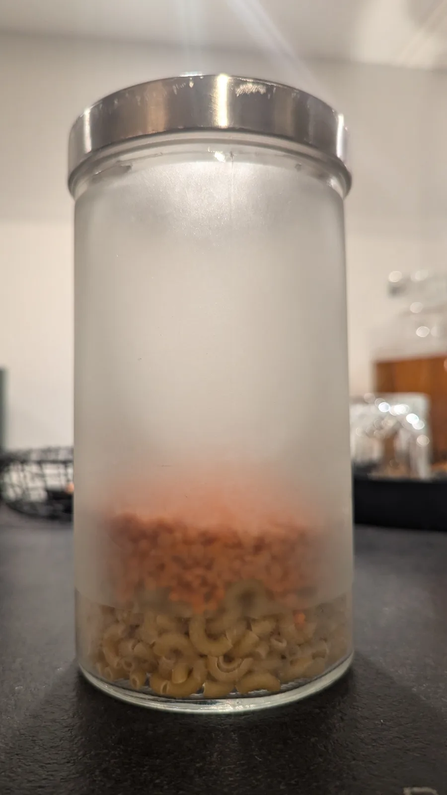 A clear glass jar with a silver lid sits on a dark countertop, containing visible layers: uncooked elbow macaroni at the…
