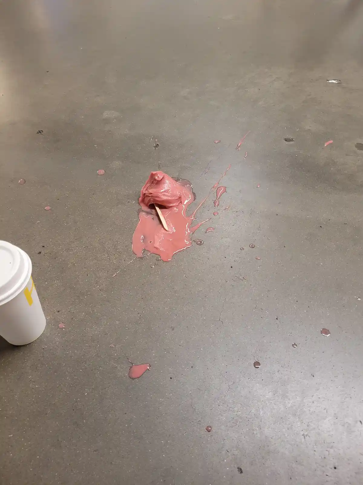 A pink, viscous substance, likely lip gloss, is spilled on a gray concrete floor with a wooden stick stuck in it, surrounded…
