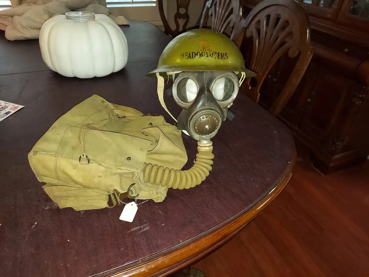 A vintage olive-drab gas mask with a matching carrying case sits on a dark wooden dining table, next to a white decorative…