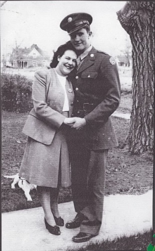 A vintage black-and-white photo shows a smiling woman in a coat embracing a man in a military uniform; they stand outdoors…
