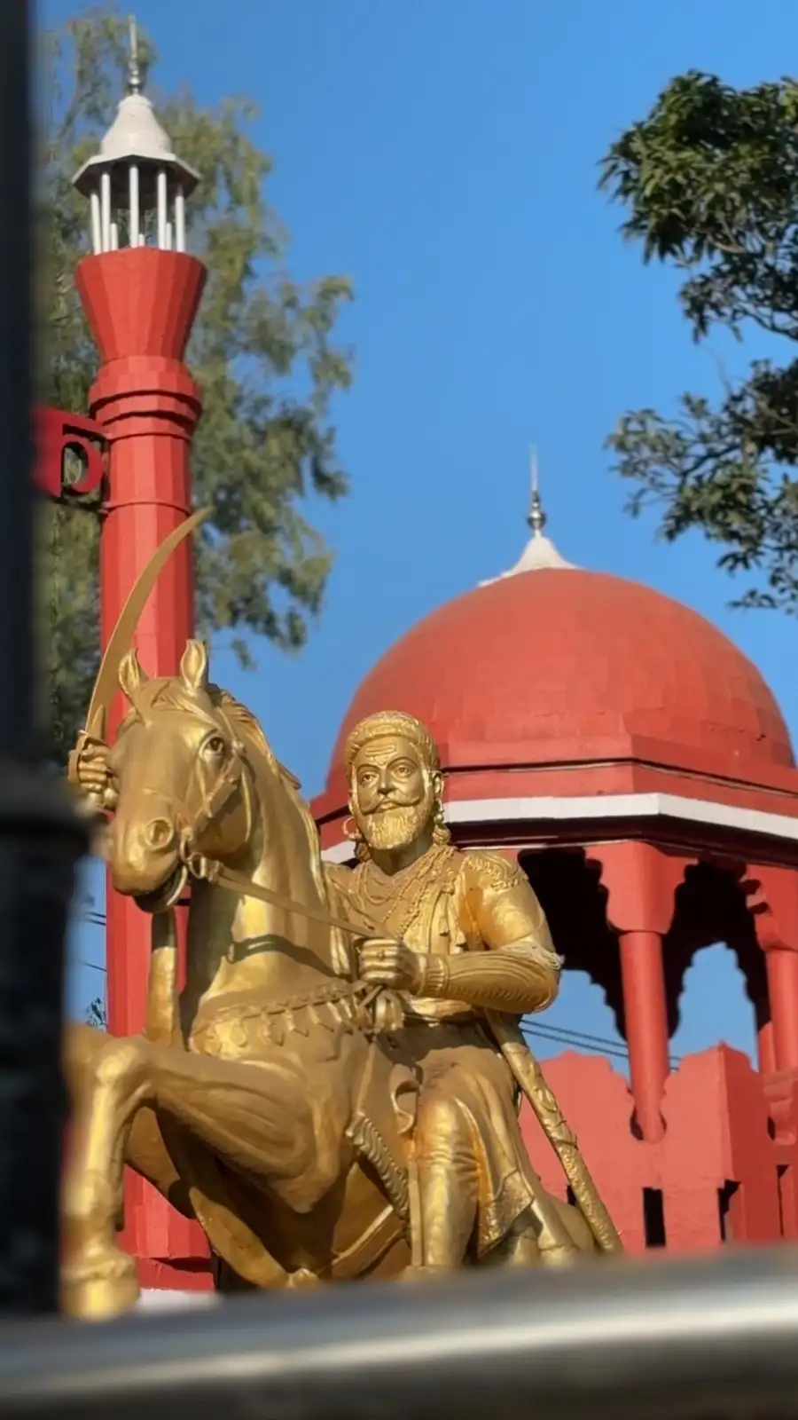 A golden statue of a bearded man on horseback holding a sword, set against a red domed building and a red pole under a clear…