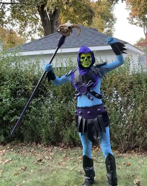 A person in a full-body costume resembling the character Skeletor from Masters of the Universe stands outdoors in a yard…