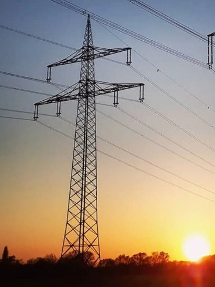 A tall, metal electricity pylon stands silhouetted against a sunset sky with orange and blue gradients. Multiple power lines…
