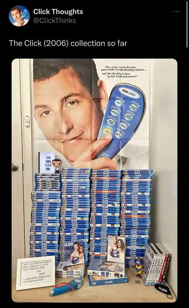 A large collection of 'Click' (2006) DVDs is stacked on a table in front of a movie poster featuring the main character…