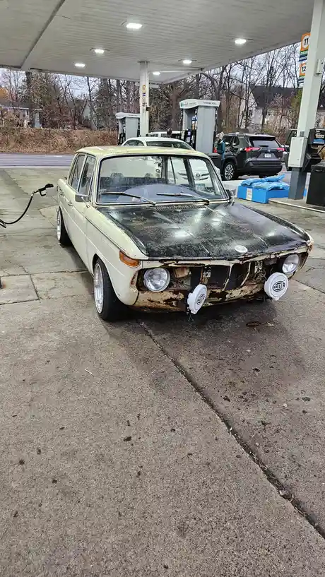 A heavily rusted, vintage white BMW sedan is parked at a gas station pump, its front bumper missing and headlights exposed…