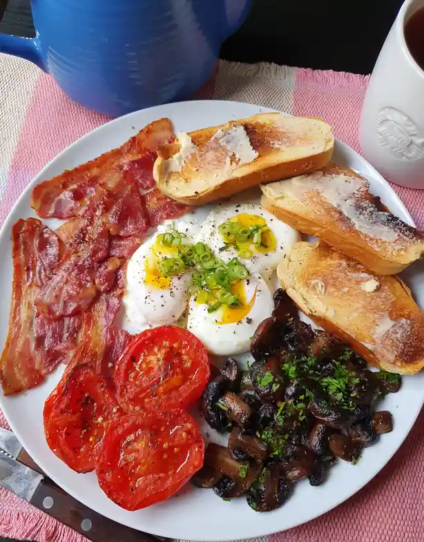 A plate of breakfast food including bacon, poached eggs with green onions, buttered toast, roasted tomatoes, and sautéed…