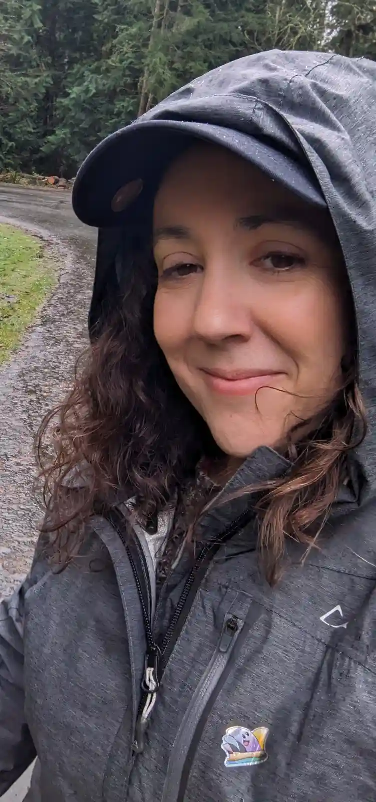 A woman with dark curly hair smiles at the camera while wearing a grey hooded rain jacket with the hood up, standing on a…