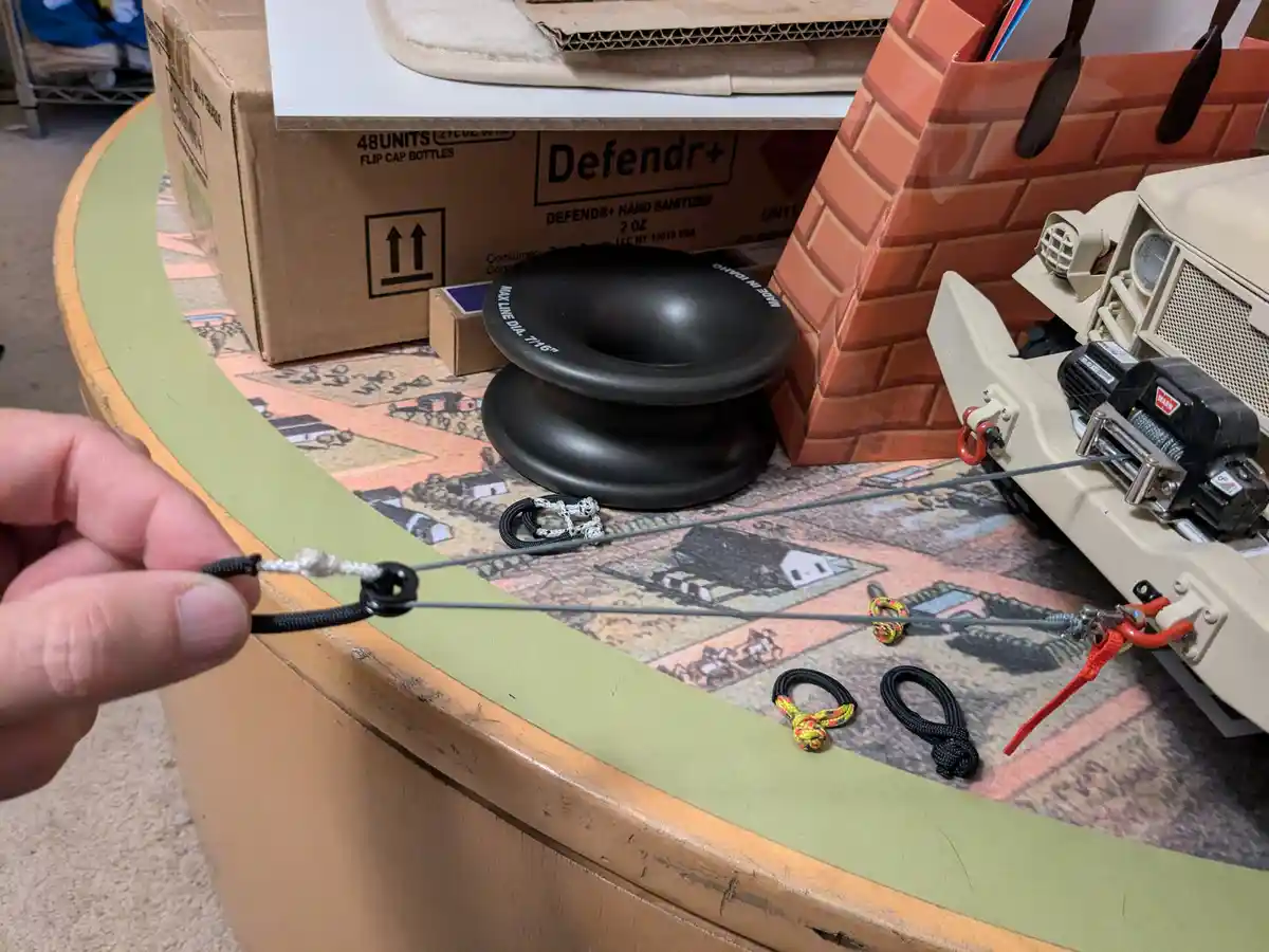 A hand holds a shoelace tied to a miniature winch cable, which is connected to a toy truck's winch. Several other shoelace…