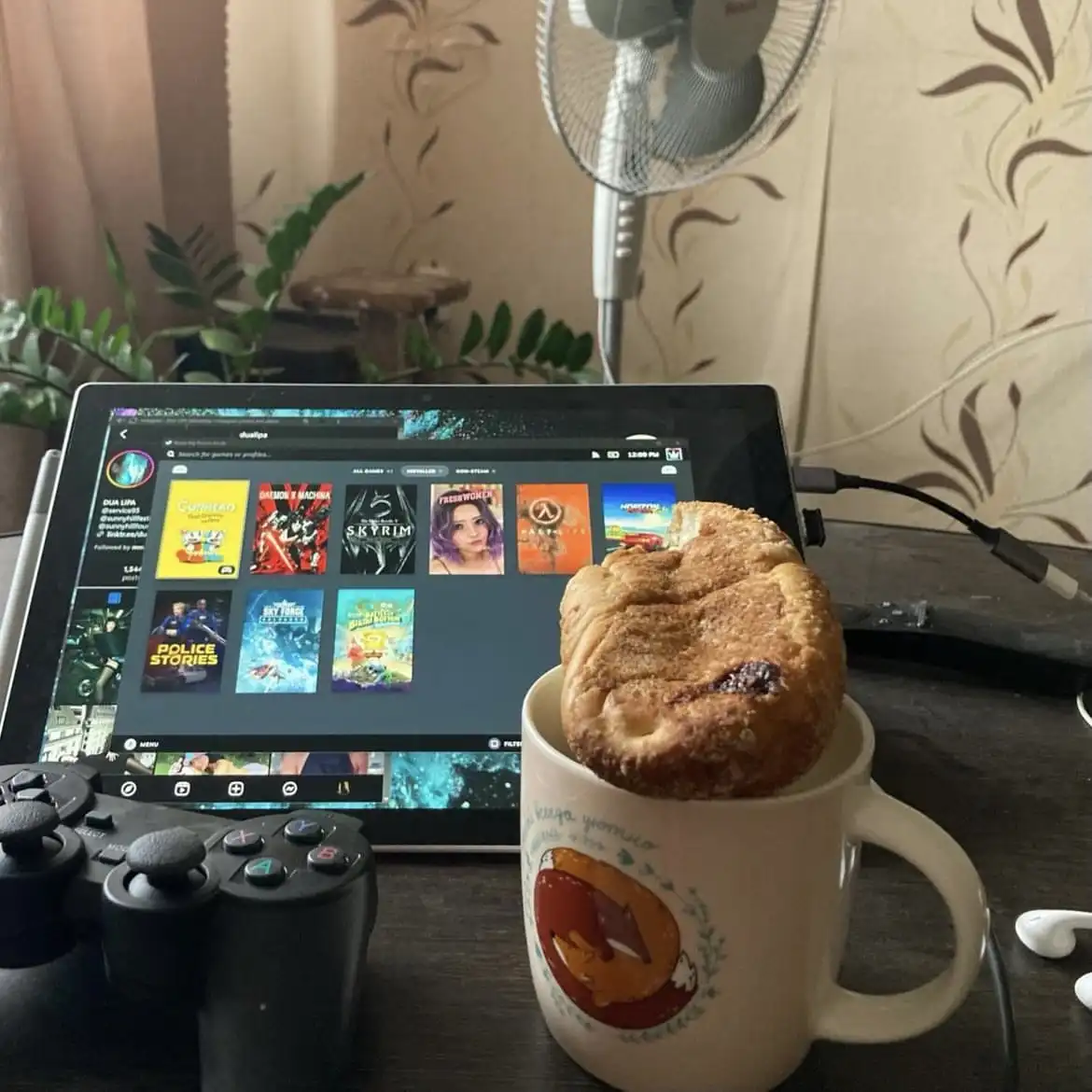 A cozy gaming setup with a tablet displaying a game library, a PlayStation controller, and a mug holding a large, sugary…