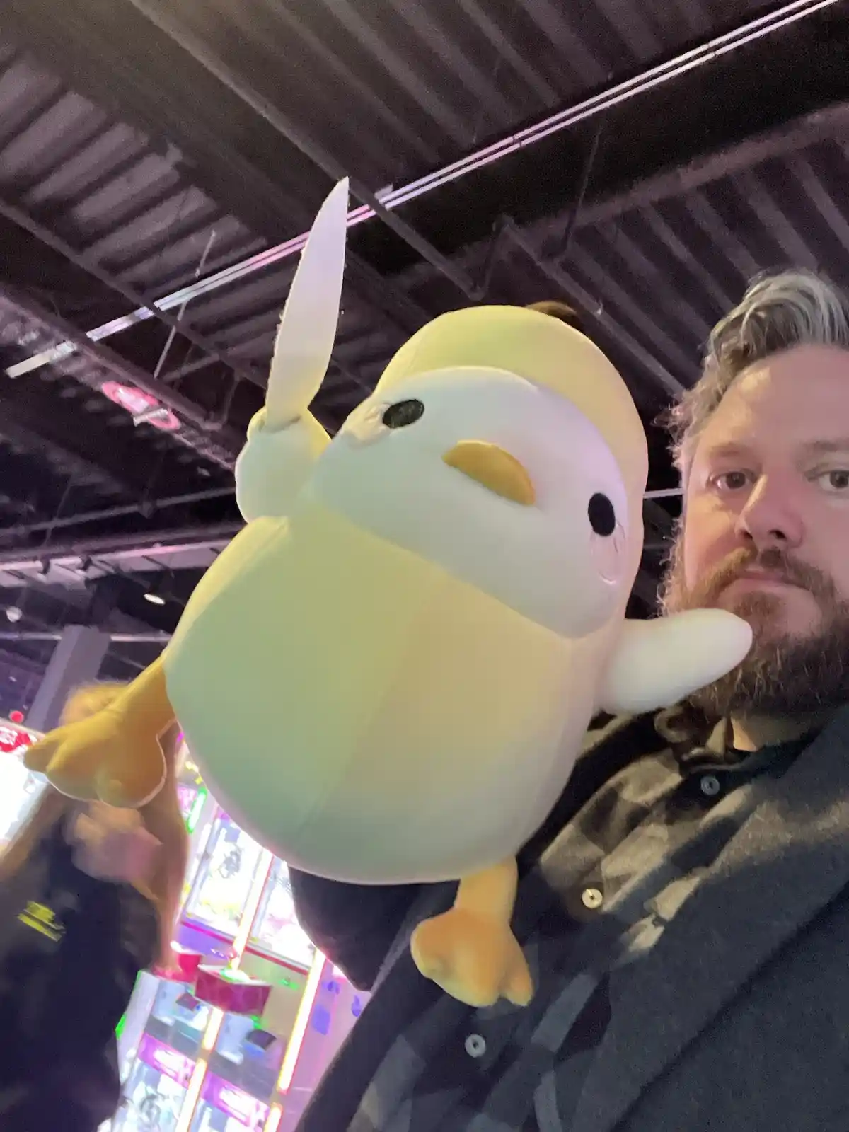A man with a beard and grey hair holds a large, yellow, plush toy resembling a bird with a white beak and a single white…