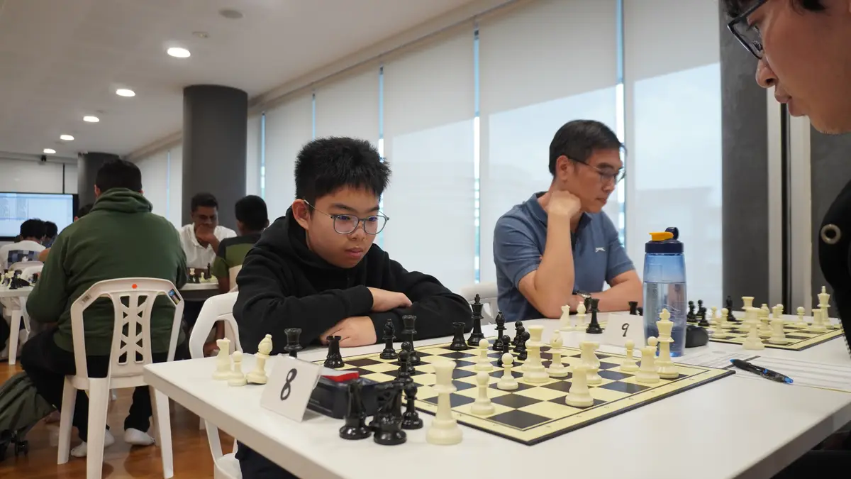 A young boy with glasses sits at a chessboard, looking intensely focused during a tournament, with other players visible in…