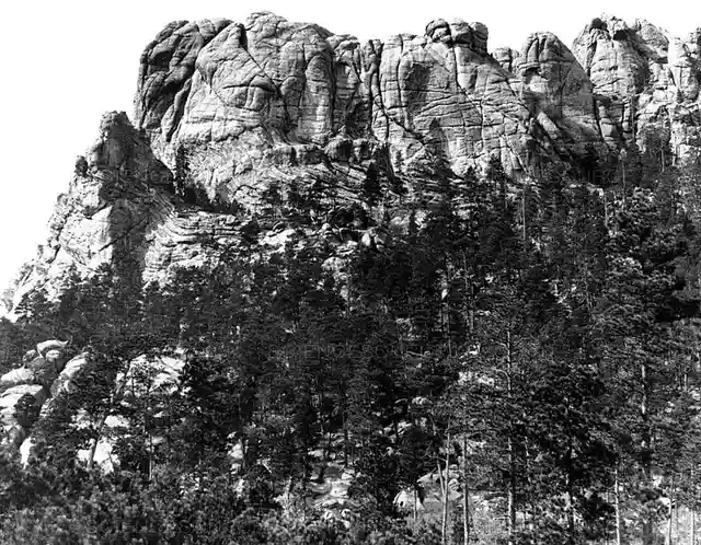 A black-and-white photograph of a rugged, rocky mountain face with dense pine trees at its base. Overlaid text in a simple…