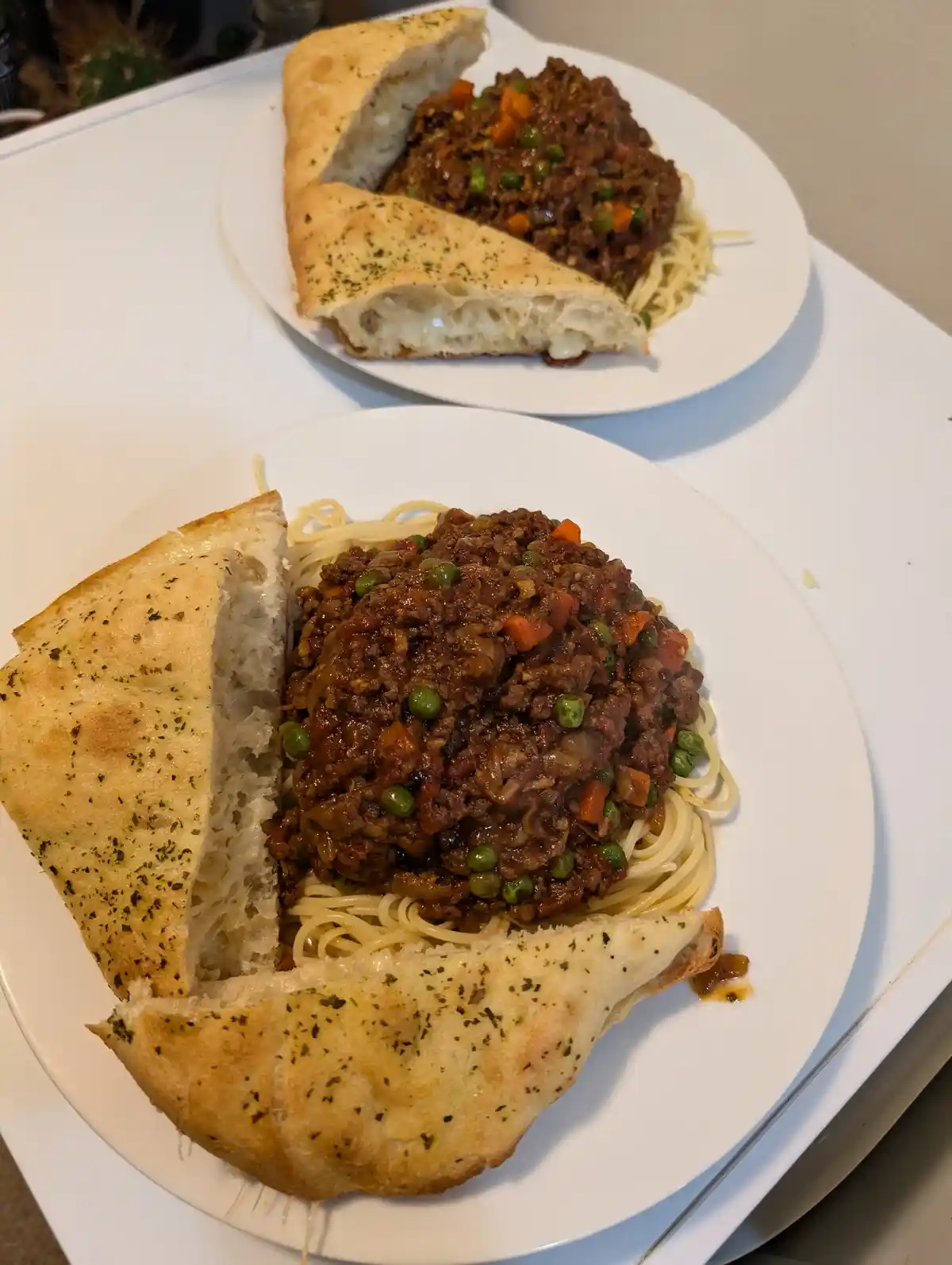 Two white plates show pasta topped with meat sauce and peas, surrounded by large pieces of garlic bread that form a…