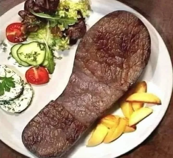 a white plate holds a large, seared steak shaped exactly like a human shoe, accompanied by a side salad with tomatoes…