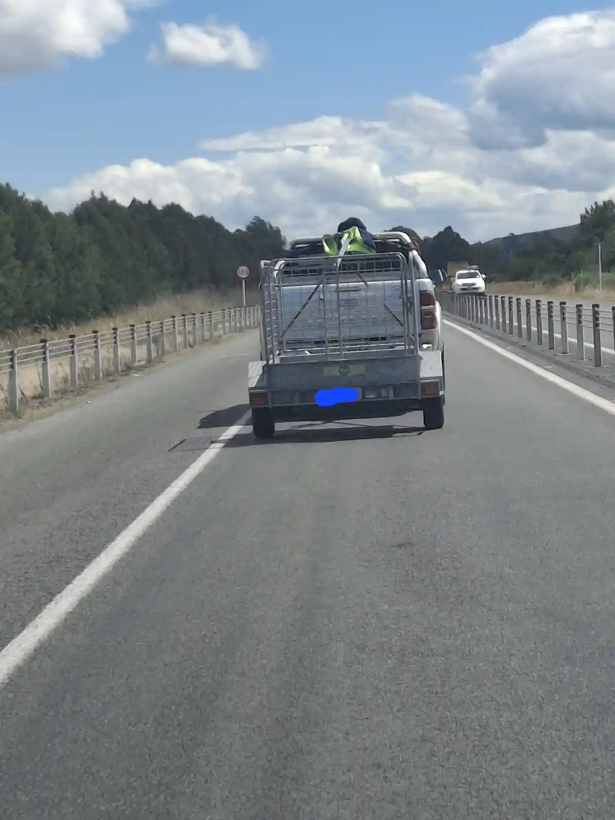 A silver pickup truck with a metal cargo trailer is driving on a highway under a partly cloudy sky. The truck's license…