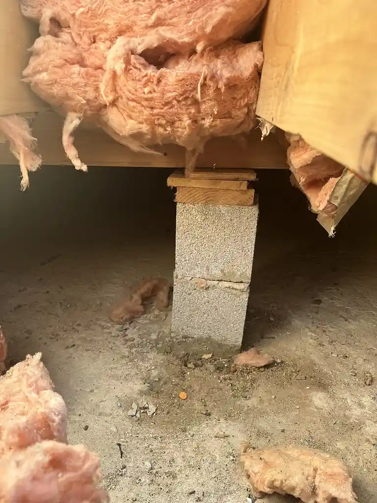 A close-up photo shows pink fiberglass insulation hanging loosely from a wooden floor joist, with some strands falling onto…