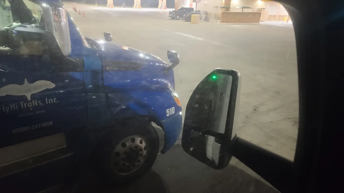 A nighttime photo taken from inside a vehicle shows a blue semi-truck parked at a rest stop. The truck's side displays…