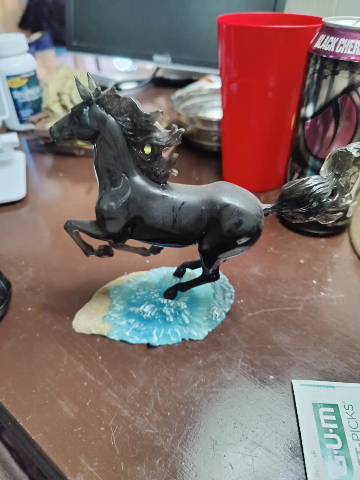 A black plastic horse statue, posed mid-gallop on a blue and beige base, sits on a cluttered desk next to a red cup, a black…