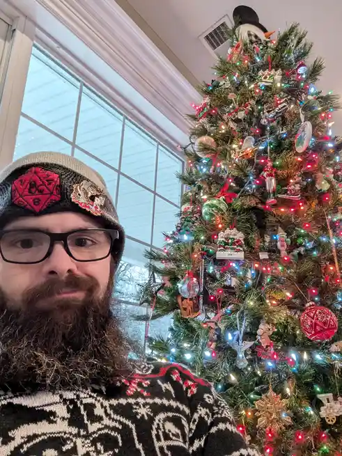 A man with a beard and glasses, wearing a beanie with a red d20 die patch and a black-and-white patterned sweater, poses for…