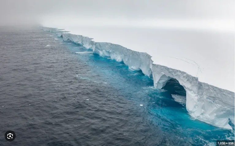 an aerial view of a massive glacier extending into dark ocean water, featuring a prominent natural archway carved through…