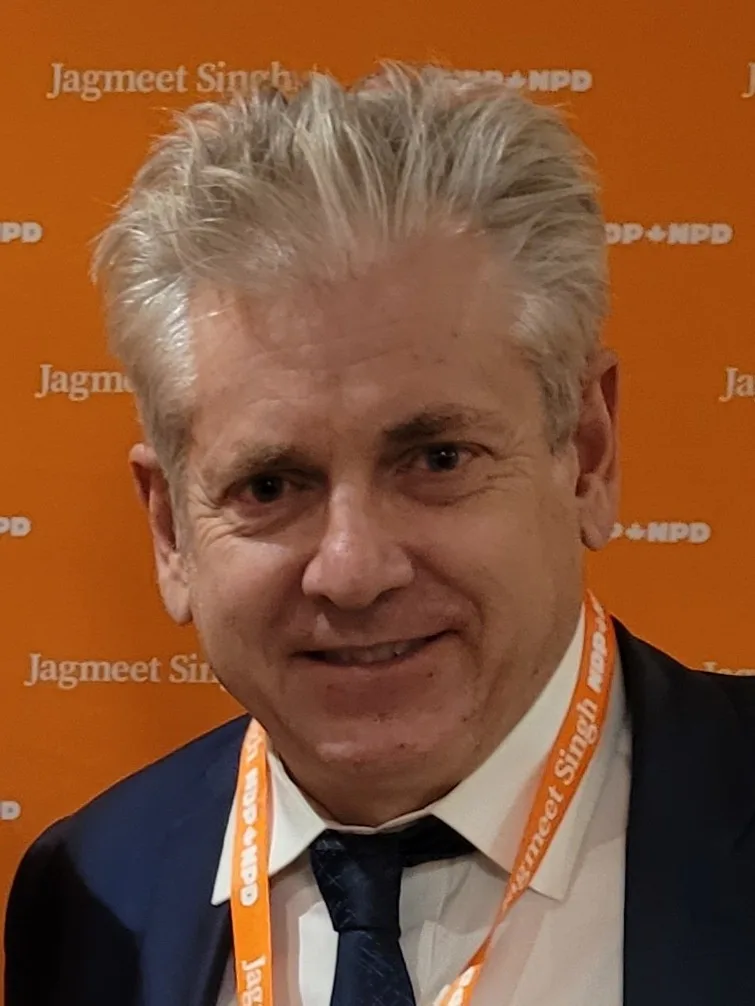 A man with gray hair wearing a suit and tie stands in front of an orange backdrop repeatedly printed with 'Jagmeet Singh…