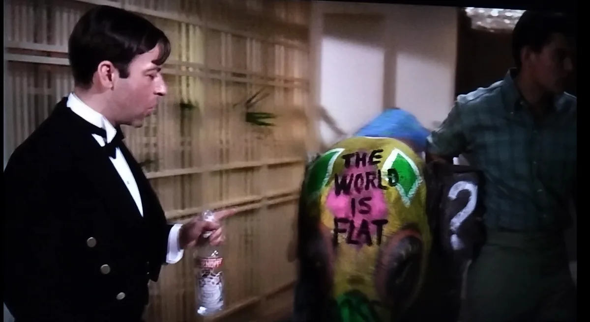 A man in a tuxedo points at another man whose back is painted with the text 'THE WORLD IS FLAT' and a large question mark on…