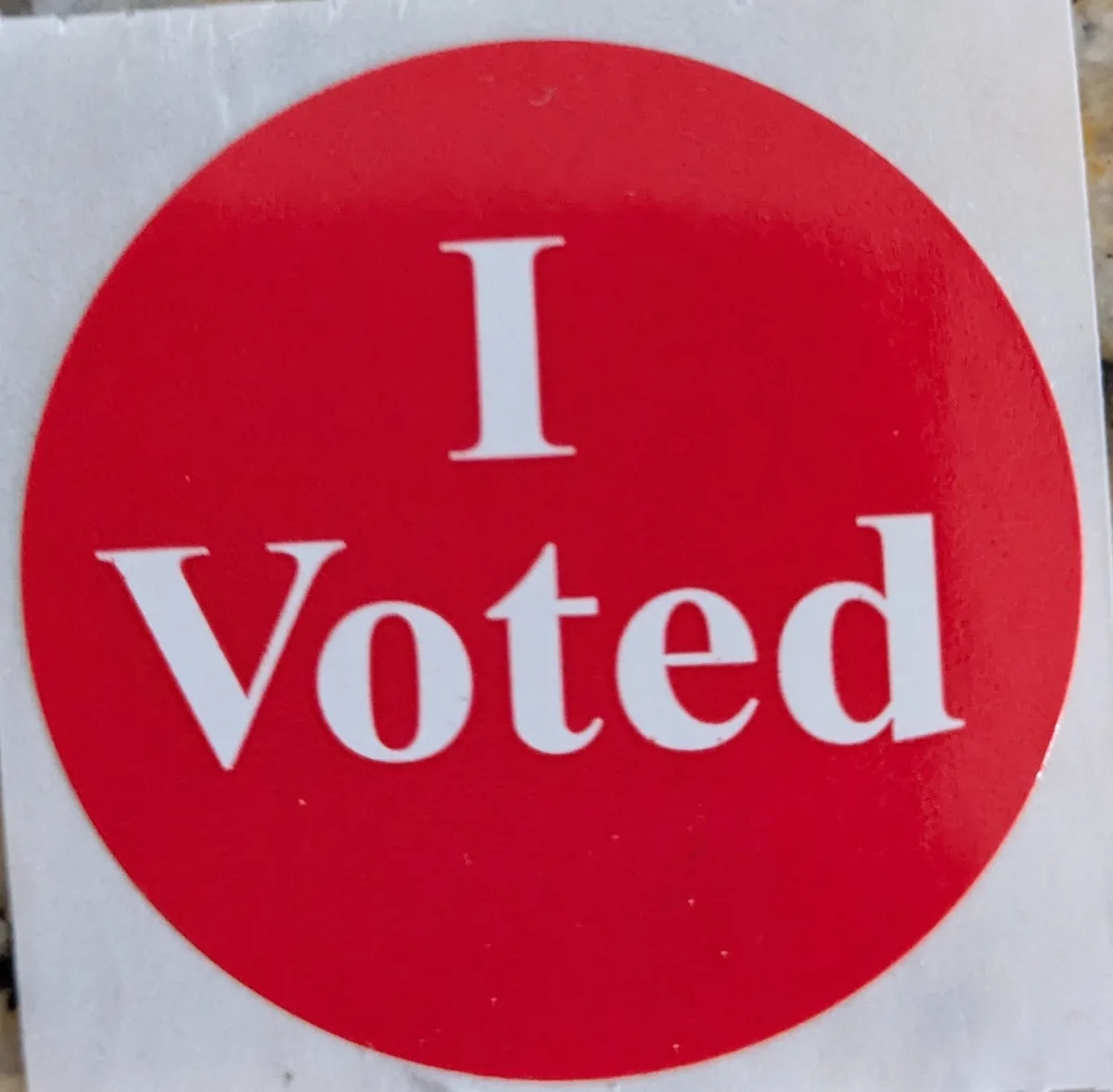 A close-up photo shows a circular red sticker with white text that reads 'I Voted', commonly given to voters after casting…