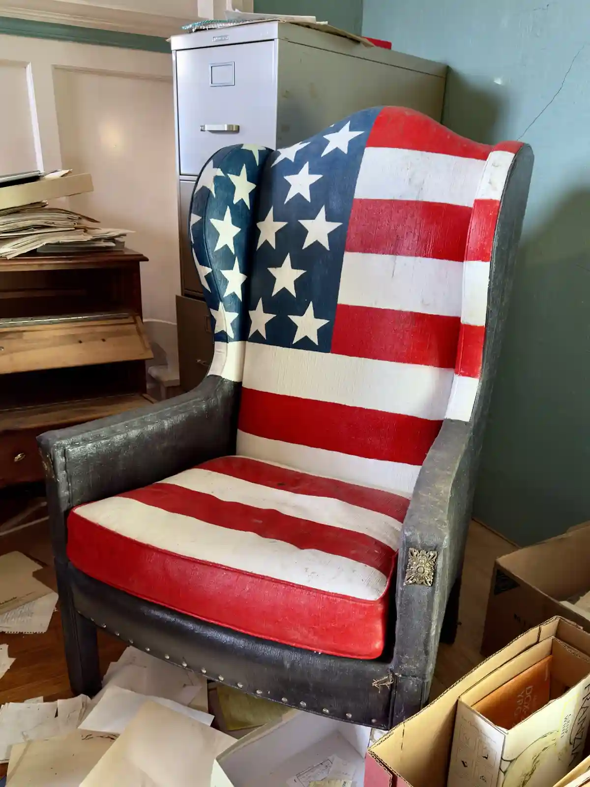 A worn, wingback armchair is painted with the American flag design, featuring stars and stripes on its back and seat, set in…