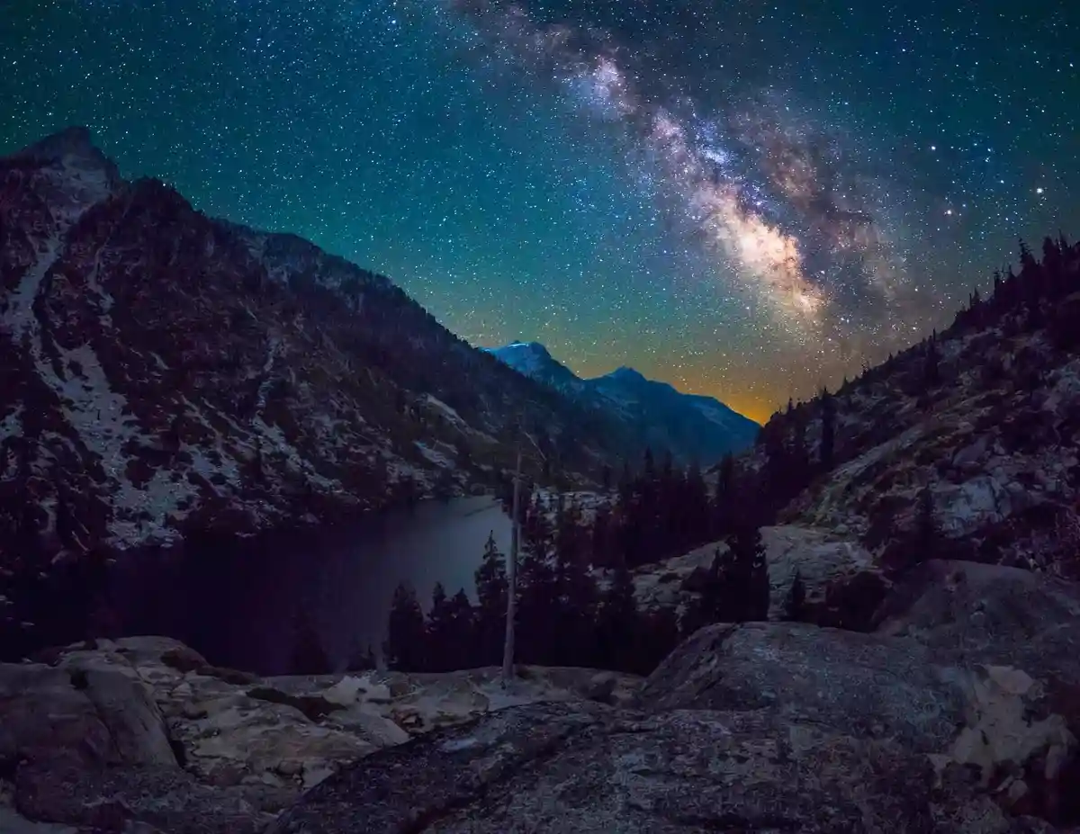 A breathtaking night photograph shows a valley with mountains, a lake, and trees under a starry sky with the Milky Way…