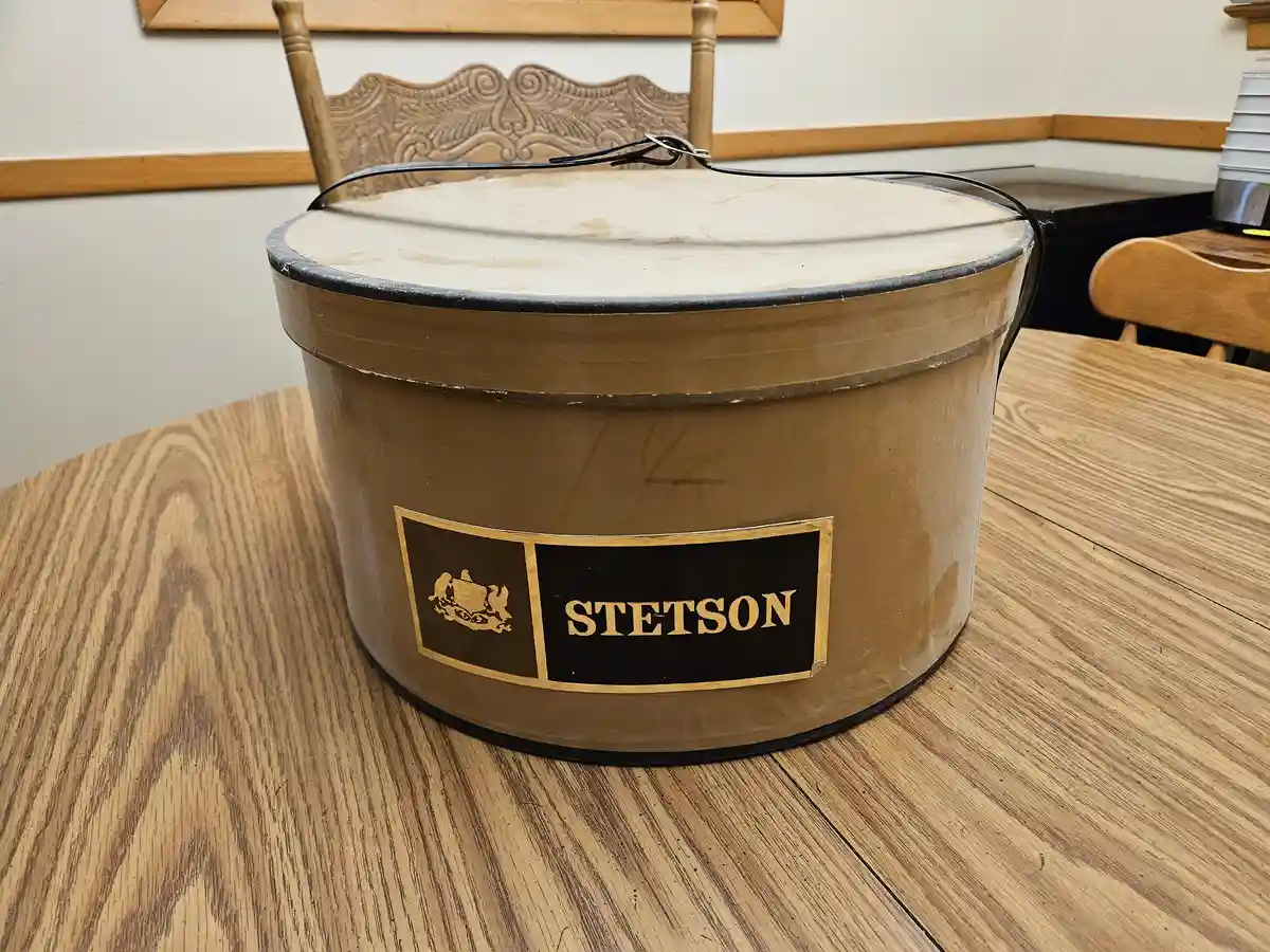 A vintage, brown, cylindrical Stetson hat box with a black and gold label sits on a wooden table. The box has a black wire…