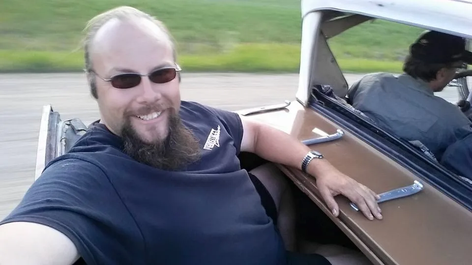 A man with a beard and sunglasses smiles while taking a selfie from the passenger side of a moving vintage car, his arm…